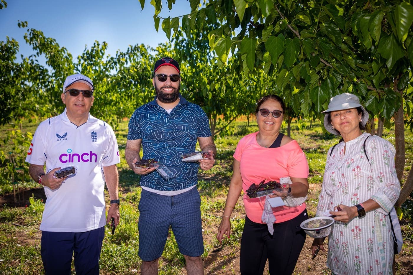 Dos parejas sostienen envases de moras en el huerto, un hombre lleva una camiseta de cricket