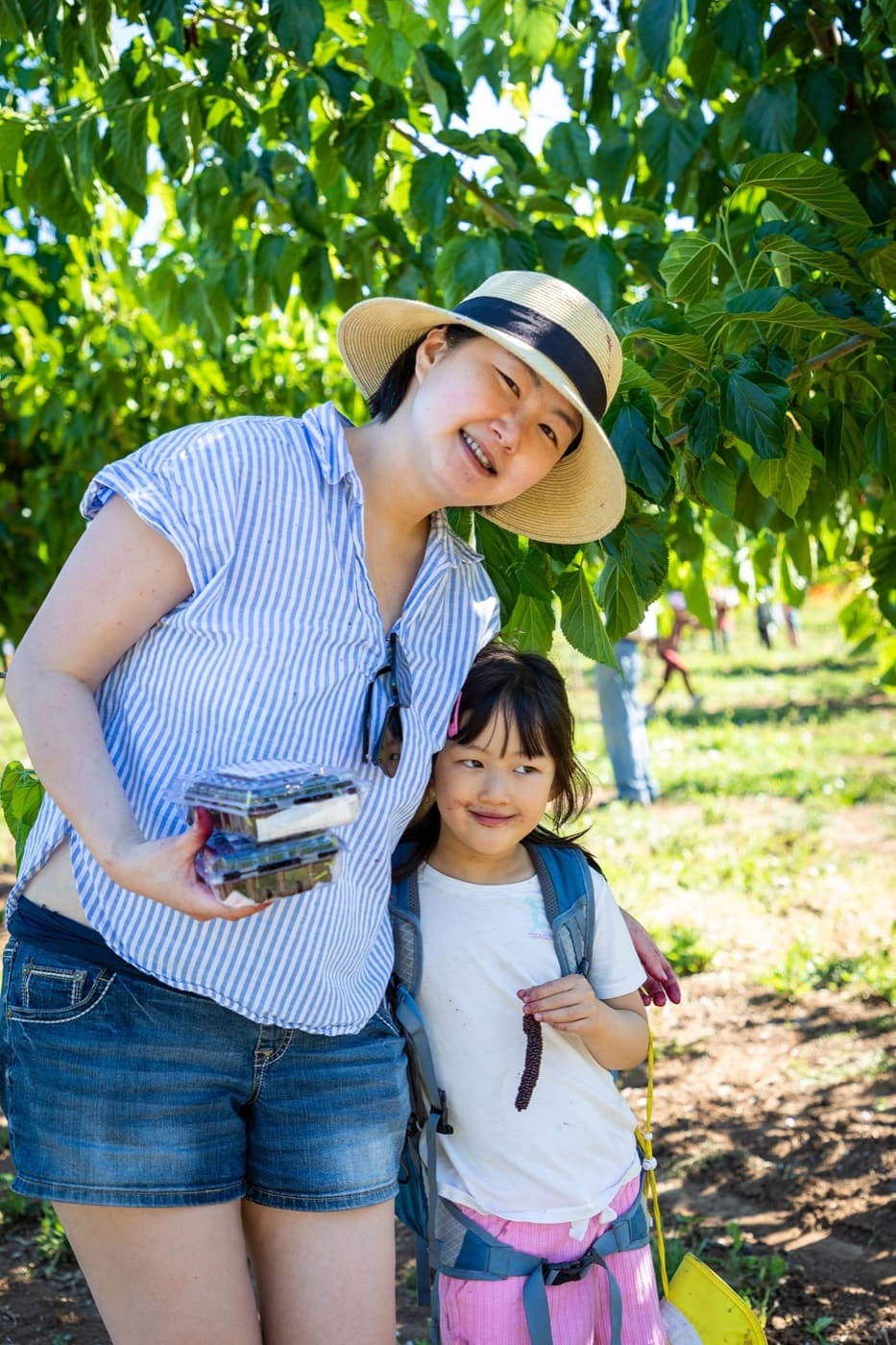 Una mamá con sombrero de paja está de pie junto a su hija, quien lleva una mochila y sostiene una rama de moras