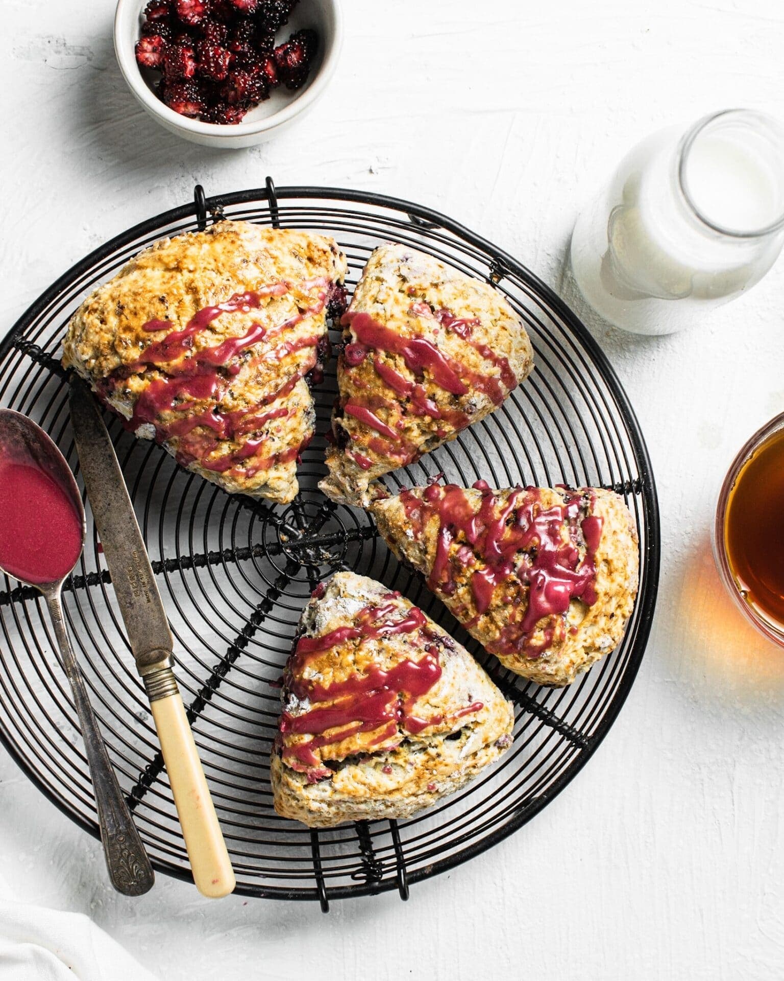 Scones triangulares de mora bañados con glaseado rosa, sobre una rejilla redonda, con mermelada, leche y té cerca, Brentwood, CA