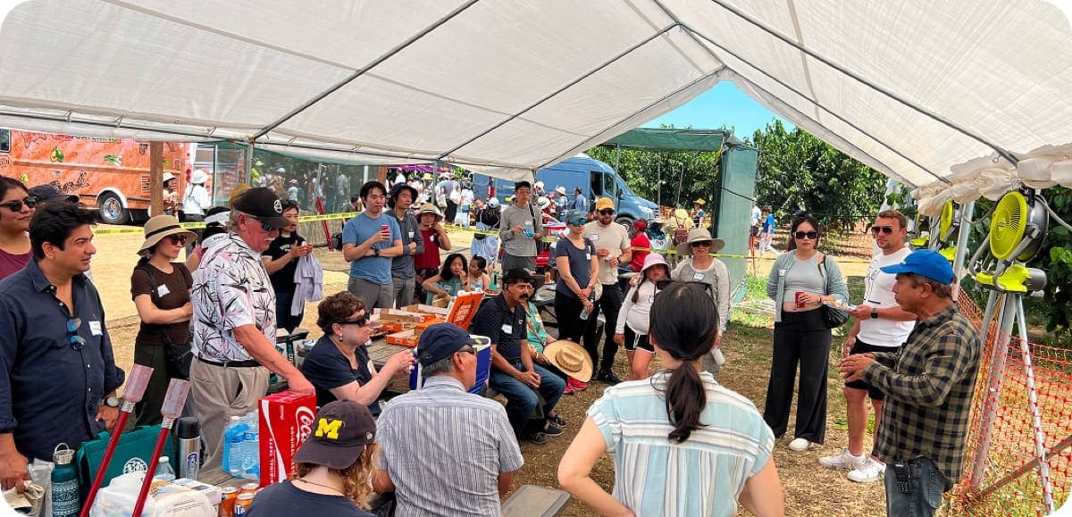 Una gran multitud reunida bajo una carpa de eventos en una reunión en la granja, con un food truck y el huerto visible afuera